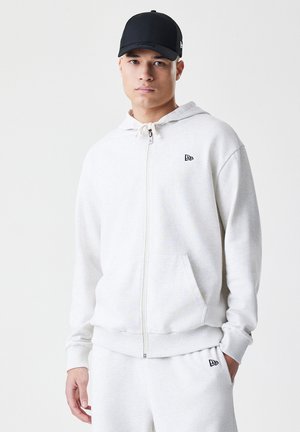 Light gray zip-up hoodie made of soft fabric, featuring a front pocket and a white drawstring. Black cap completes the look.