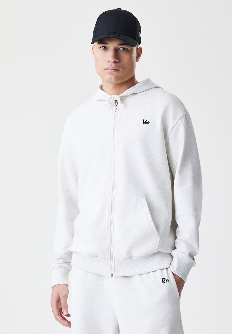 Lys grå zip-up hoodie lavet af blødt stof, med en frontlomme og en hvid snor. En sort kasket fuldender looket.