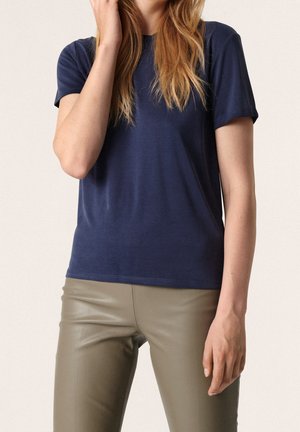 Woman wearing a navy blue short-sleeve t-shirt and olive green leather pants, with long blonde hair partially visible.