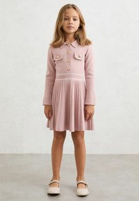 Rochie plisată roz cu mâneci lungi, guler cu nasturi și două buzunare la piept. Are cusături albe și pantofi bej cu funde, cu talpă plată.