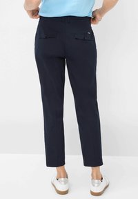 Marineblaue Hosen mit geradem Schnitt, ausgestattet mit zwei Gesäßtaschen mit Klappen und Knöpfen. Getragen mit weißen Schuhen und einem hellblauen Oberteil.