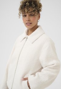 White zip-up jacket with a textured fabric, wide collar, and oversized fit. Features side pockets and a soft, cozy appearance.