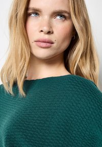 Blonde Frau mit blauen Augen und leichten Sommersprossen trägt einen strukturierten dunkelgrünen Pullover und schaut leicht nach oben und zur Seite.