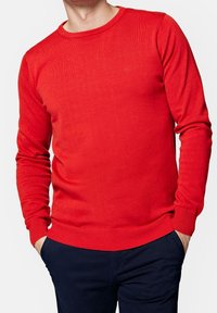 Pull en tricot rouge en matériau doux, avec un col rond et des manches longues, doté de bords côtelés aux poignets et à l'ourlet. Petit logo sur la poitrine.