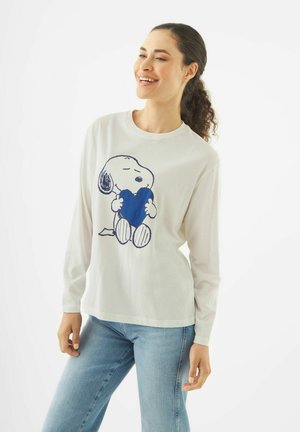 Junge Frau lächelt, trägt ein weißes Langarmshirt mit Snoopy, der ein blaues Herz hält, kombiniert mit hellblauen Jeans, stehend vor weißem Hintergrund.