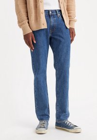 Person som bär blå denimjeans, blå canvas sneakers, en vit skjorta och en beige stickad kavaj mot en enfärgad bakgrund.