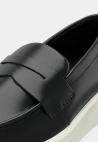 Μαύρο δερμάτινο μοκασίνι με λουράκι για πέννι και λευκή σόλα από καουτσούκ, εμφανίζοντας κοντινή λήψη της επάνω και πλευρικής ραφής.