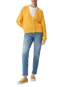 Keltainen neulottu neuletakki, jossa on V-kaula-aukko, nappisulku ja väljä malli; yhdistettynä vaaleansinisiin kuluneisiin farkkuihin ja oransseihin tennareihin.