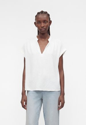 Γυναίκα με πλεξούδες που φοράει λευκό μπλουζάκι με κοντά μανίκια και V-λαιμόκοψη και ανοιχτά μπλε τζιν, στέκεται μπροστά σε φόντο απλό.