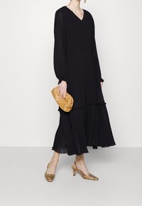 Abito lungo a maniche lunghe nera con scollatura a V, dettagli arricciati, abbinato a una pochette beige e scarpe slingback con motivo dorato.