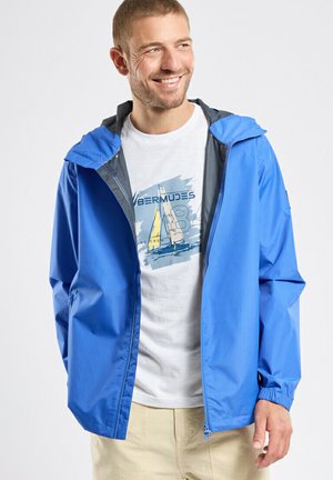 Homme souriant portant une veste à capuche bleue sur un T-shirt blanc à thème nautique et un pantalon beige, debout devant un fond clair uni.