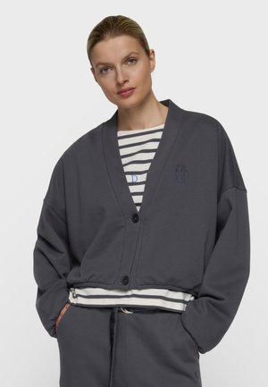 Blonde Frau, die eine dunkelgraue, geknöpfte Strickjacke über einem schwarz-weiß gestreiften Hemd und passenden dunkelgrauen Hosen trägt, Hände in den Taschen.