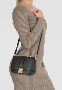 Borsa in pelle nera con una chiusura dorata, forma strutturata, manico corto e tracolla removibile, indossata con un vestito beige a costine.