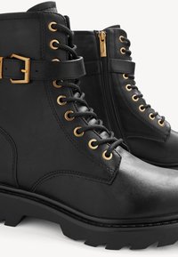 Bottes de combat en cuir noir avec œillets dorés, fermeture éclair latérale, lacets à l'avant et détail de sangle à boucle sur des semelles robustes.