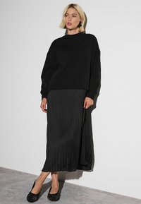 Black oversized sweatshirt paired with a pleated, ankle-length black skirt and textured black flats. Smooth fabric with subtle sheen.