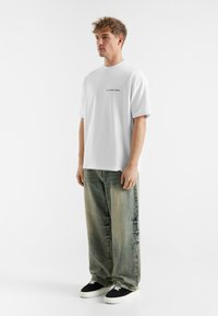 Witte katoenen T-shirt met zwarte tekst op de voorkant, gecombineerd met losse, vervaagde blauw-groene denim broeken en zwarte sneakers.