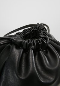 Pochette noire en cuir avec cordon de serrage, dessus froncé et fine sangle sur un fond clair uni.