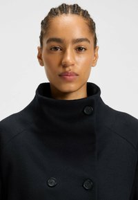 Donna con capelli intrecciati indossa un cappotto nero doppiopetto con colletto alto su uno sfondo semplice chiaro.