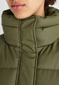Veste matelassée vert olive avec un col montant, présentant des coutures horizontales, une texture lisse et un petit détail avec une fermeture éclair noire.