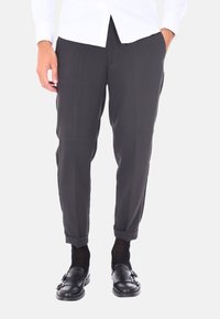 Pantaloni neri su misura con taglio dritto, orli arrotolati, realizzati in un tessuto liscio. Indossati con mocassini neri e calze scure.