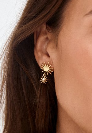 Pendientes dorados en forma de sol con un colgante de sol más pequeño colgando debajo, en una persona con cabello largo y marrón y piel marrón clara.