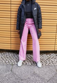 Abrigo acolchado negro, suéter de cuello alto negro, pantalones de pierna ancha rosa claro y zapatillas blancas. Presenta un logo de Nike en el abrigo y en la camiseta. Fondo texturizado.