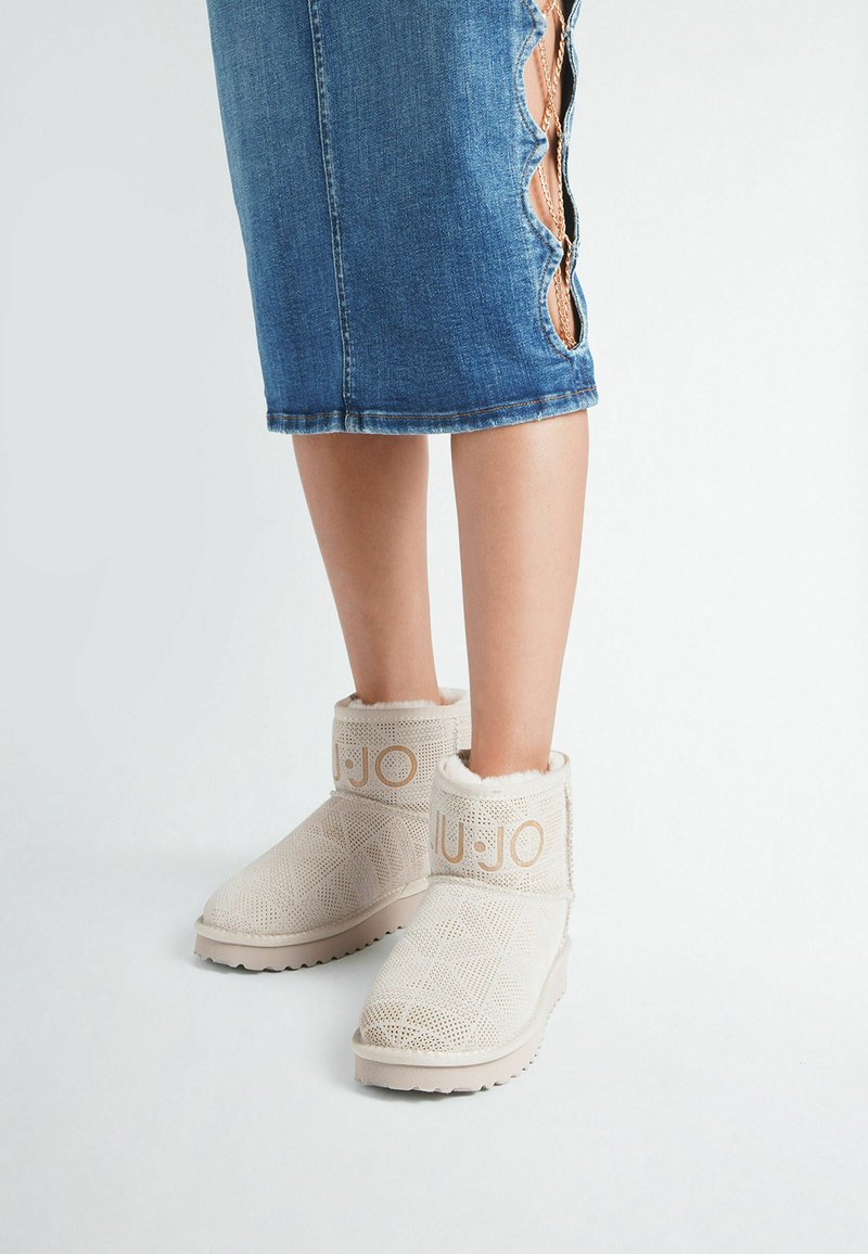 Beine, die einen blauen Jeansrock mit gewellten Schlitzen und beigen, mit Pelz gefütterten Knöchelstiefeln mit strukturiertem Muster und "LIU·JO"-Markierung tragen.