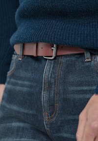 Donkerblauwe jeans met oranje stiksels, een bruine leren riem met een metalen gesp, en een gestructureerde marineblauwe gebreide trui.