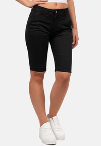 Bermuda shorts en denim noir avec cinq poches, une fermeture éclair, et une silhouette ajustée, portés avec des baskets blanches de sport.