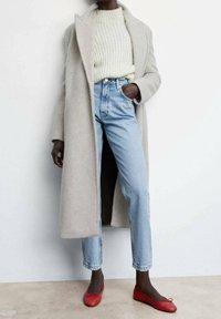 Personne portant un long manteau beige clair, un pull en maille crème, un jean taille haute bleu clair, et des ballerines rouges, debout à l'intérieur.