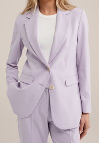 Kvinna som bär en ljuslila kostymjacka och matchande byxor med en vit ribbad t-shirt, hand i fickan på jackan.