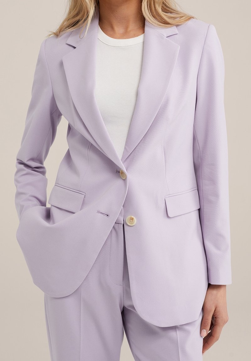Kvinna som bär en ljuslila kostymjacka och matchande byxor med en vit ribbad t-shirt, hand i fickan på jackan.