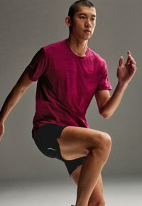 Athlète en t-shirt rouge et short noir lève un genou, effectuant un exercice dynamique sur un fond gris uni.
