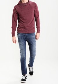 Bordeauxfärgad hoodie med huva och tryckknappdetaljer, kombinerad med slitna blåjeans och svarta sneakers med vita sulor.