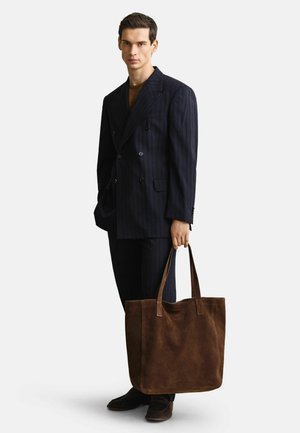 Mann in dunklem Nadelstreifenanzug und braunem Hemd, der eine große braune Wildleder-Tasche mit zwei Griffen hält.