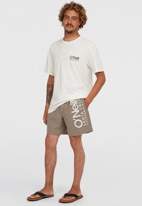 Balta kokvilnas T-kreklu ar melnu logo, bēšas peldbikses ar baltu druku un melnas flip-flops. Ikdieniška vasaras apģērbu kombinācija.