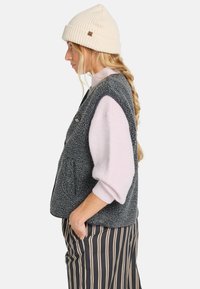 Fleece vest in donkergrijs, met een gestructureerde oppervlakte, zijzakken en een logo; gecombineerd met een lichtroze trui en gestreepte broek.