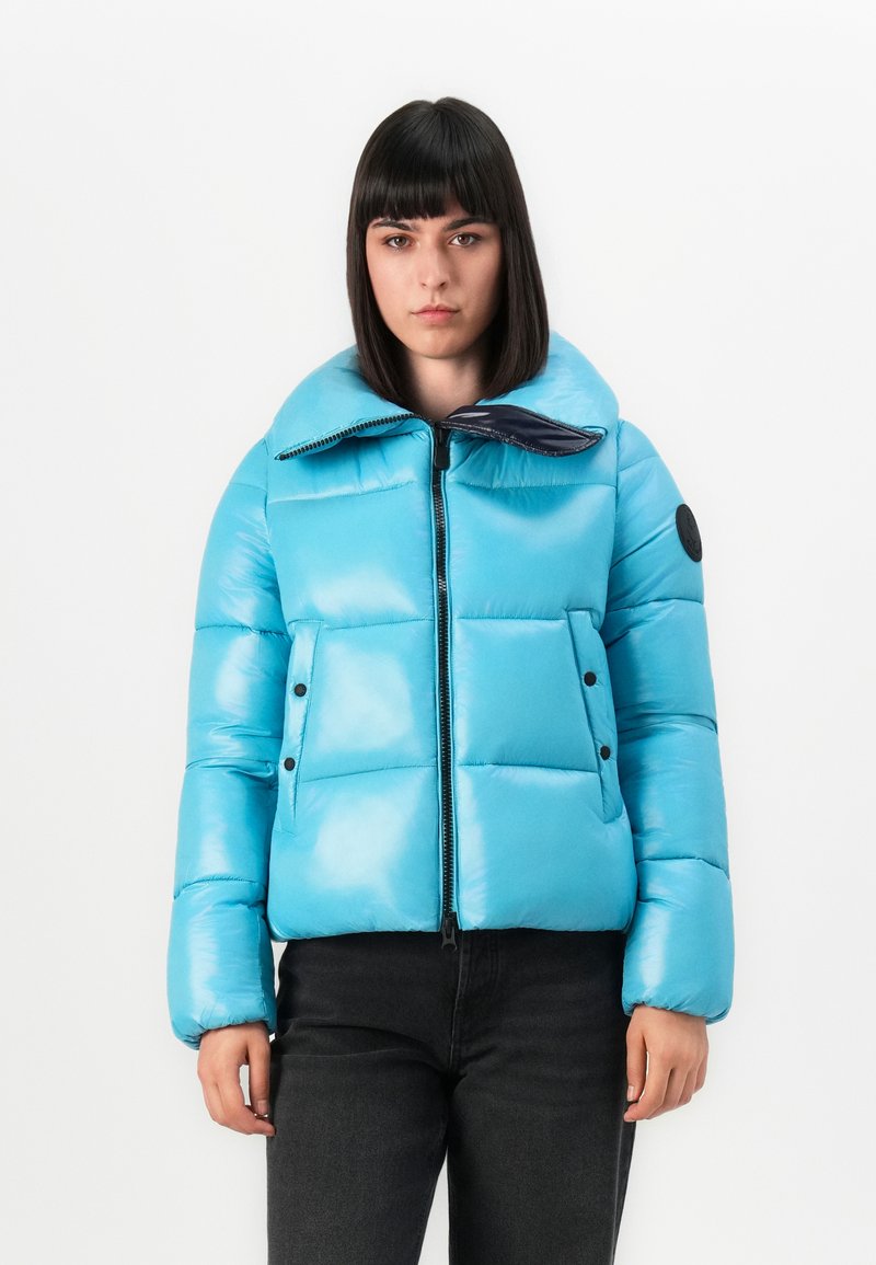 Jonge vrouw met steil, donker haar die een heldere blauwe gevoerde jas met hoge kraag en zwarte broek draagt tegen een witte achtergrond.