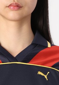 Marineblå og rød sportstrøye med kragedesign, utstyrt med gule detaljer og en liten gull Puma-logo på brystet. Teksturert stoff synlig.