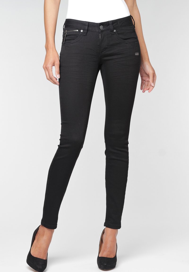 Femme portant un jean noir slim avec un détail de fermeture éclair sur les poches avant et des talons hauts noirs, debout devant un fond uni.