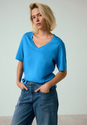 Lyshåret kvinde stående med hænderne i lommerne på jeans iført en skarp blå T-shirt med V-hals og korte ærmer mod en ensfarvet lys baggrund.