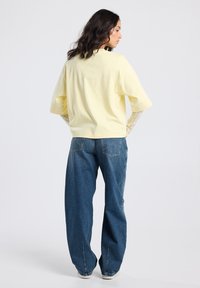 Woman with long dark hair wearing a light yellow oversized short-sleeve top over lace sleeves and wide-leg blue jeans, standing facing away.