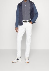 Marineblauwe doorgestikte jas over een marineblauw polkadothemd, witte broek en grijze sneakers met marineblauwe accenten. Een zwarte riem maakt de look compleet.