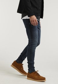 Muž kráčející v tmavých štíhlých džínách, hnědých nubukových šněrovacích botách, černé bundě a s vrstvenými černobílými košilemi na bílém pozadí.