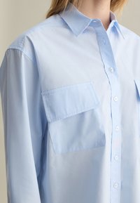 Camisa azul claro de botones en tejido suave, con cuello, dos bolsillos en el pecho y un corte recto con un ajuste relajado.