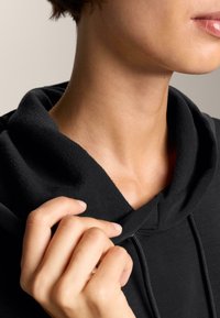 La felpa nera con cappuccio presenta un morbido rivestimento in fleece testurizzato, un cordino e una vestibilità rilassata. Una mano tira delicatamente il colletto.