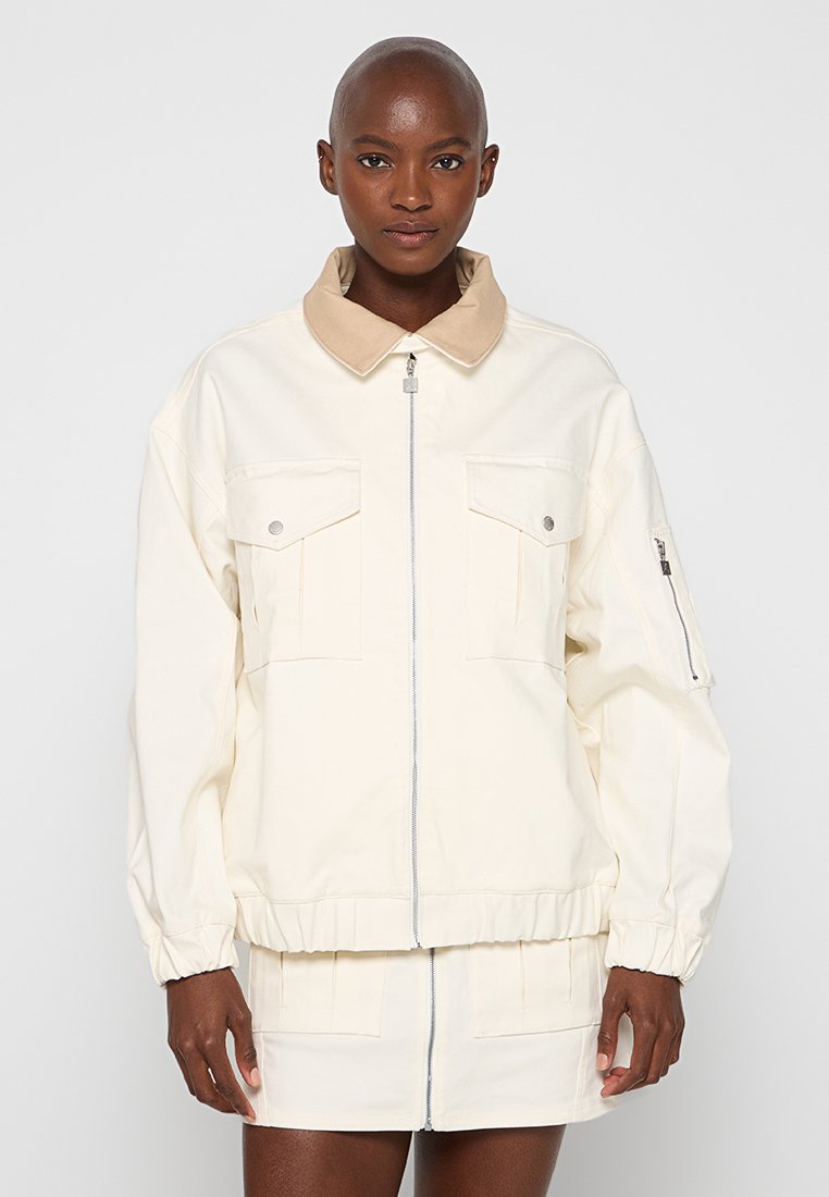 Chaqueta crema con un cuello beige, dos bolsillos frontales, puños elásticos y cierre de cremallera. Textura suave con un corte estructurado y un diseño minimalista.