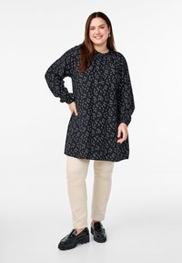 Tunique noire à motif avec des manches longues, un col rond et une patte de boutonnage, associée à un pantalon beige et des mocassins à plateforme noirs.