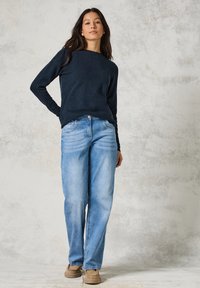 Marineblauer Pullover und hellblaue weit geschnittene Jeans, Denim mit Ausbleichdetails, kombiniert mit braunen Freizeitschuhen vor einem strukturierten hellen Hintergrund.