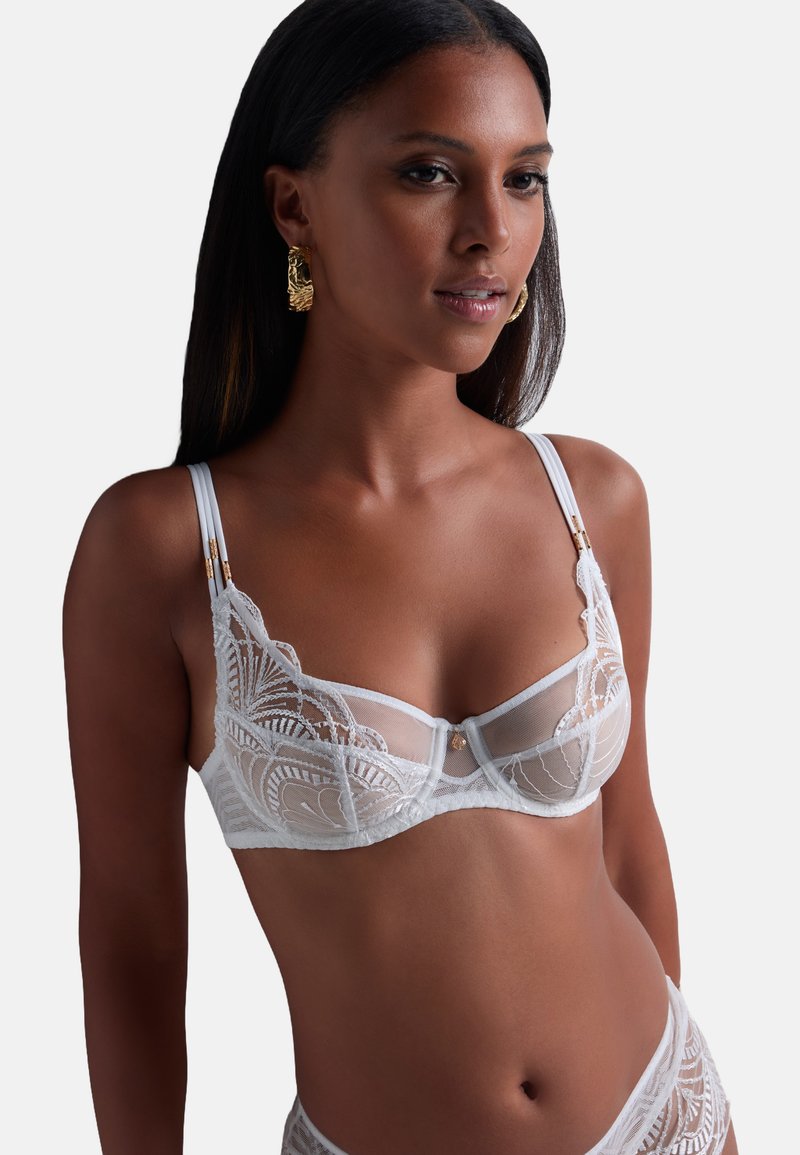 Soutien-gorge en dentelle blanche avec des panneaux transparents et une broderie complexe. Il présente des bretelles ajustables et des accents en métal doré.